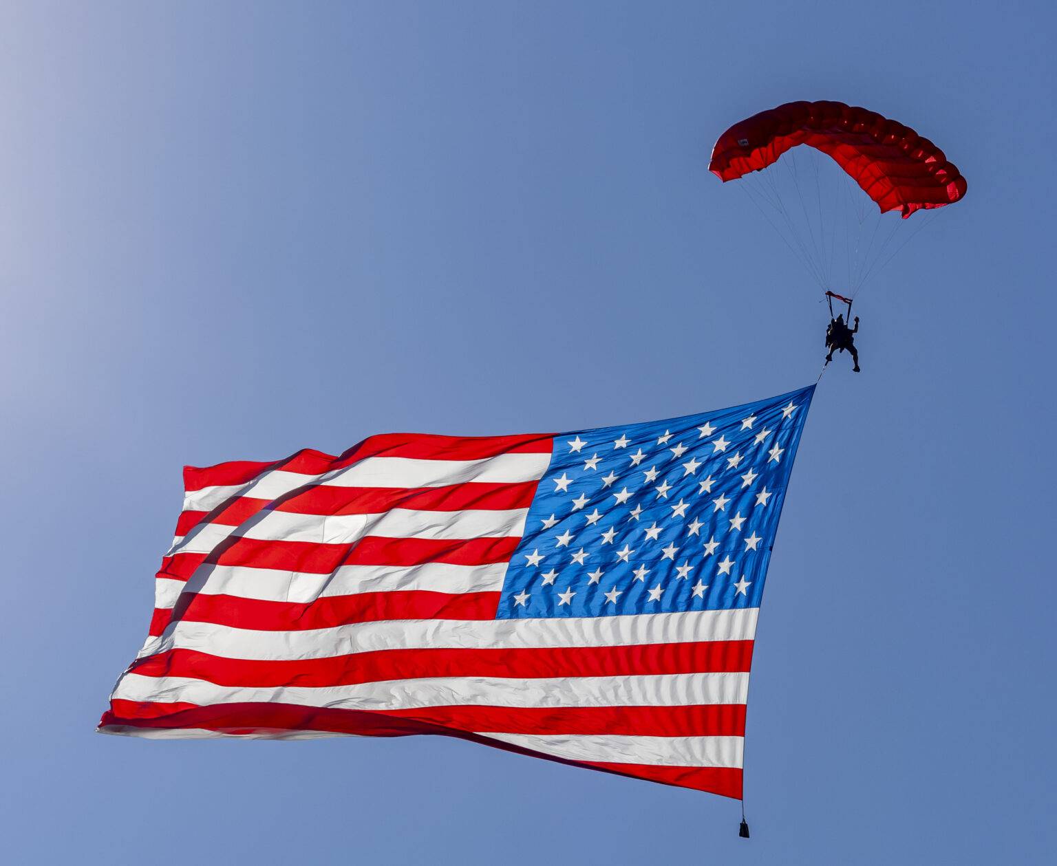 Children of Fallen Heroes Parachute Team member flying a large American flag under canopy against a clear blue sky