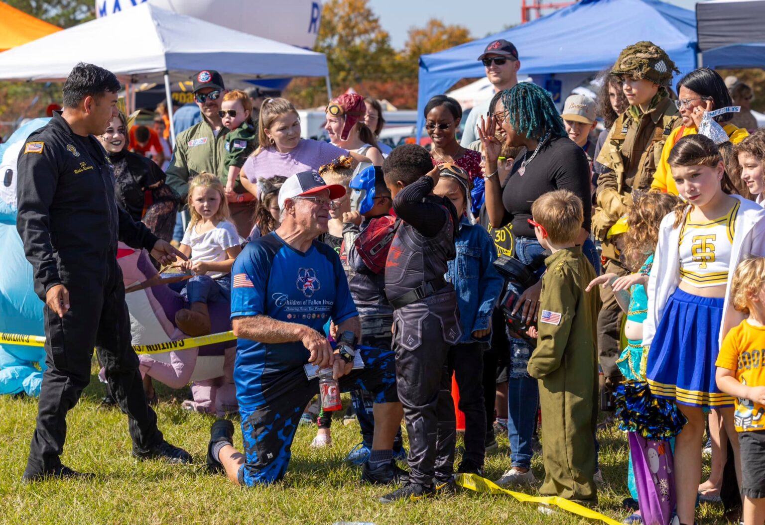 A Children of Fallen Heroes parachute team member kneeling in the grass to talk with a group of children at an outdoor community event.