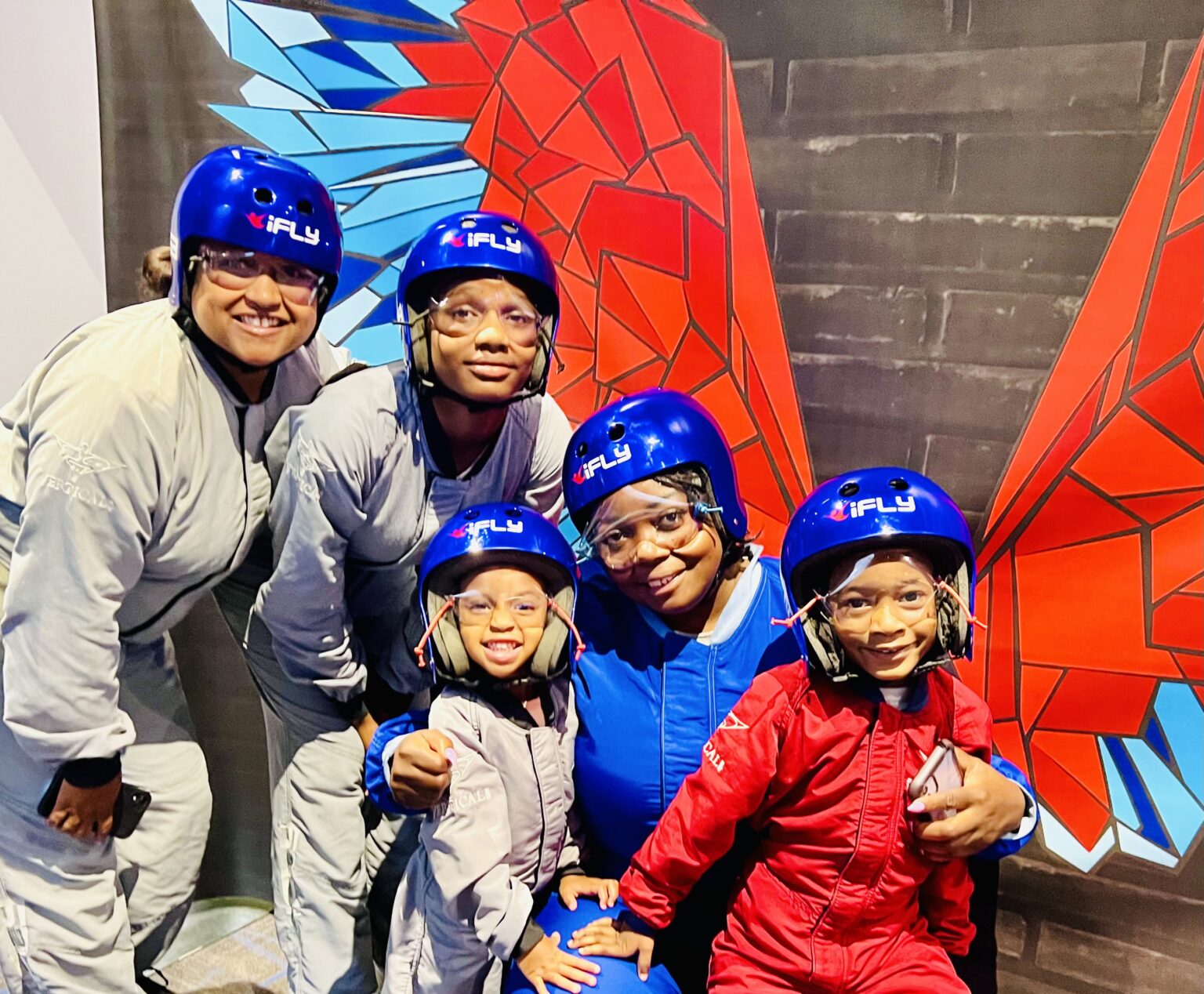 A family in flight suits and helmets smiling together before an indoor skydiving flight.