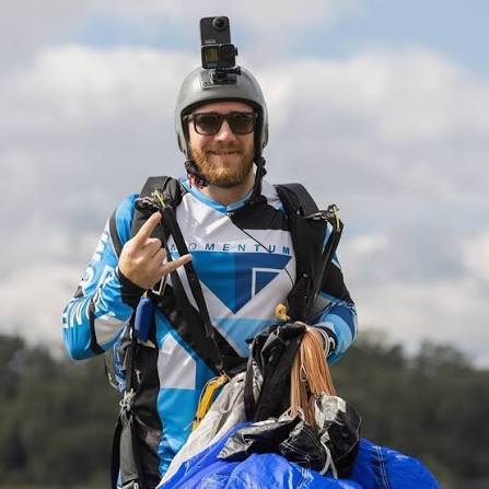 Brandon Schmidt in skydiving gear, wearing a helmet with a mounted camera and holding a packed canopy.