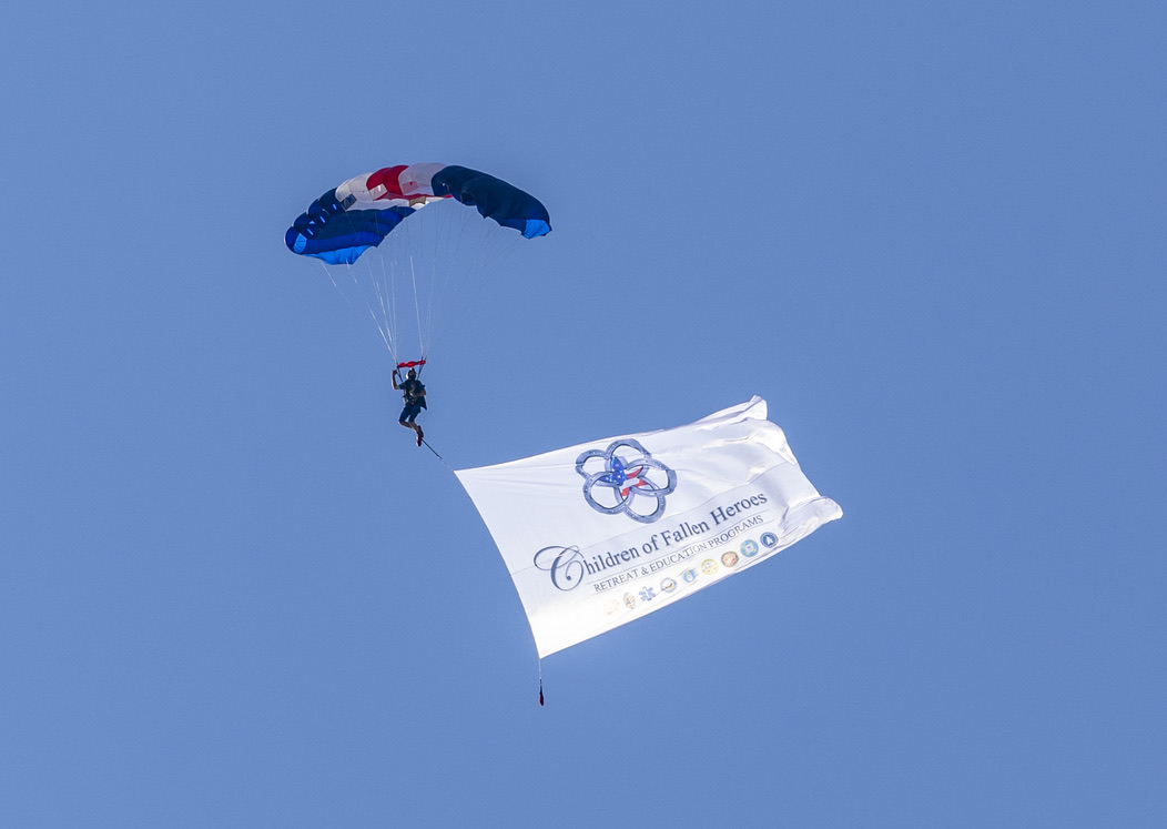 A parachutist under canopy flying the original Children of Fallen Heroes flag against a clear blue sky.