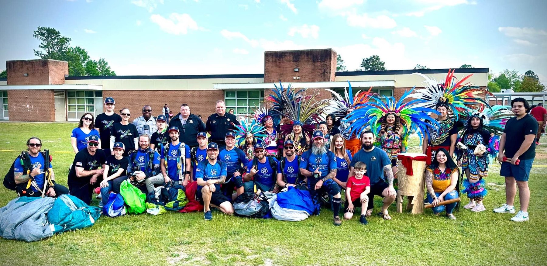Children of Fallen Heroes parachute team members with parachutes packed at their feet, first responders and Cultural Day performers in feathered regalia, gathered on the field at Cliffdale Elementary School.
