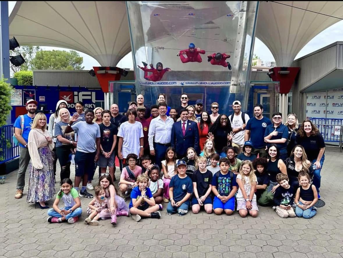 Children, families, veterans and supporters gathered for a group photograph outside the indoor skydiving venue, with flyers visible in the wind tunnel above them.