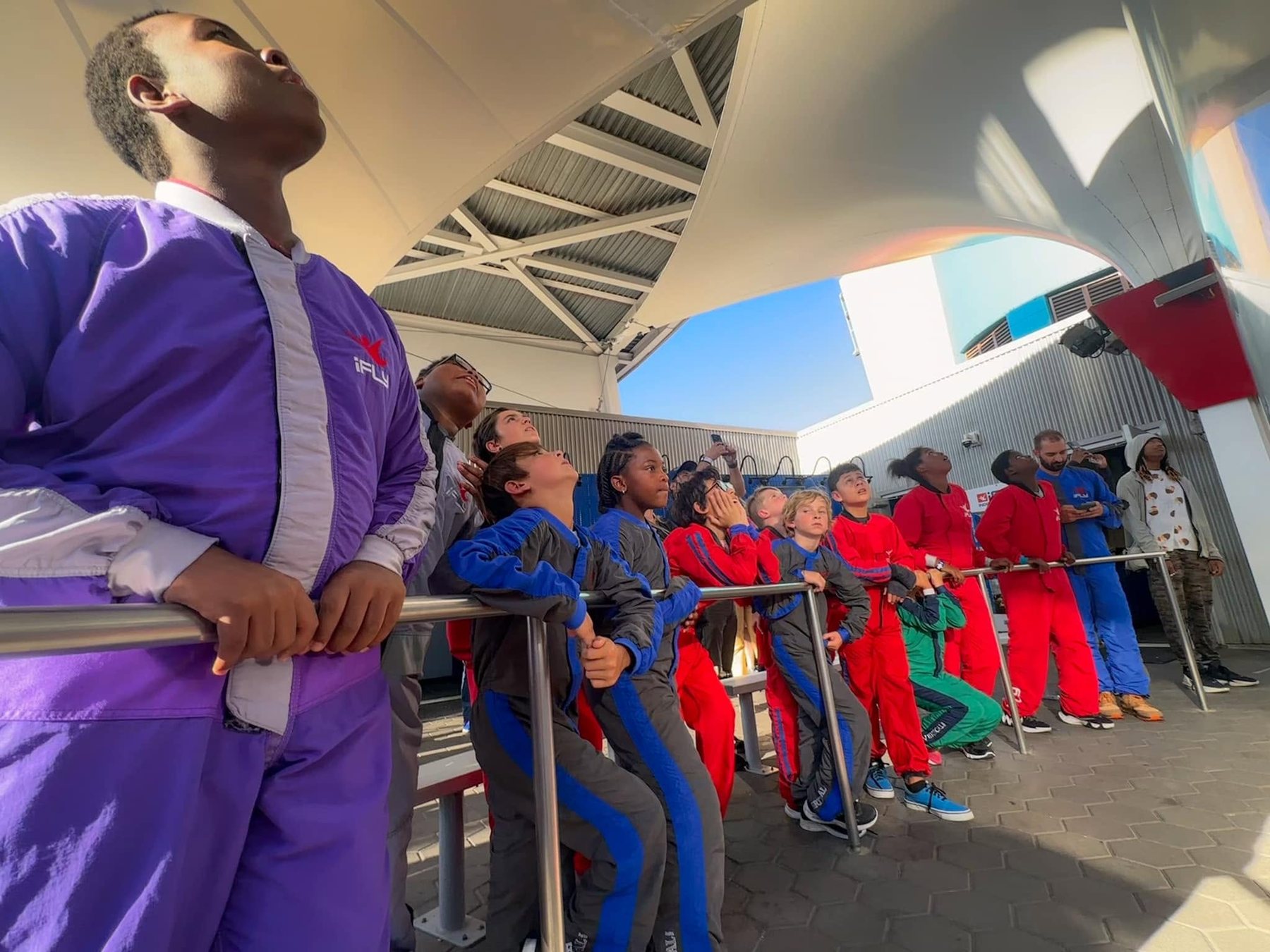 Young participants in indoor skydiving flight suits stand along a railing, looking upward as they wait for their turn in the wind tunnel.