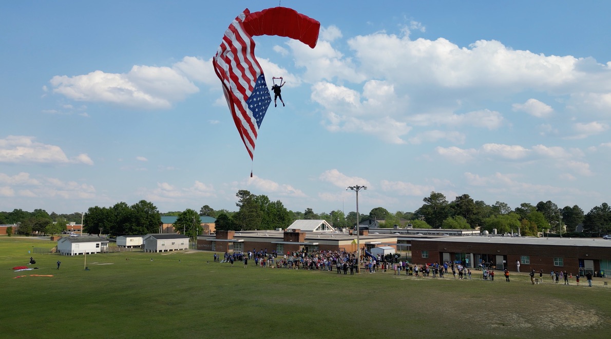A Children of Fallen Heroes parachutist descending under an American flag canopy above a school field