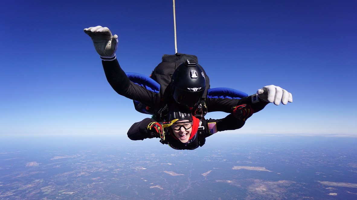 Joshua&rsquo;s twin sister Kiki in freefall on a tandem skydive, arms out and smiling, with farmland far below.