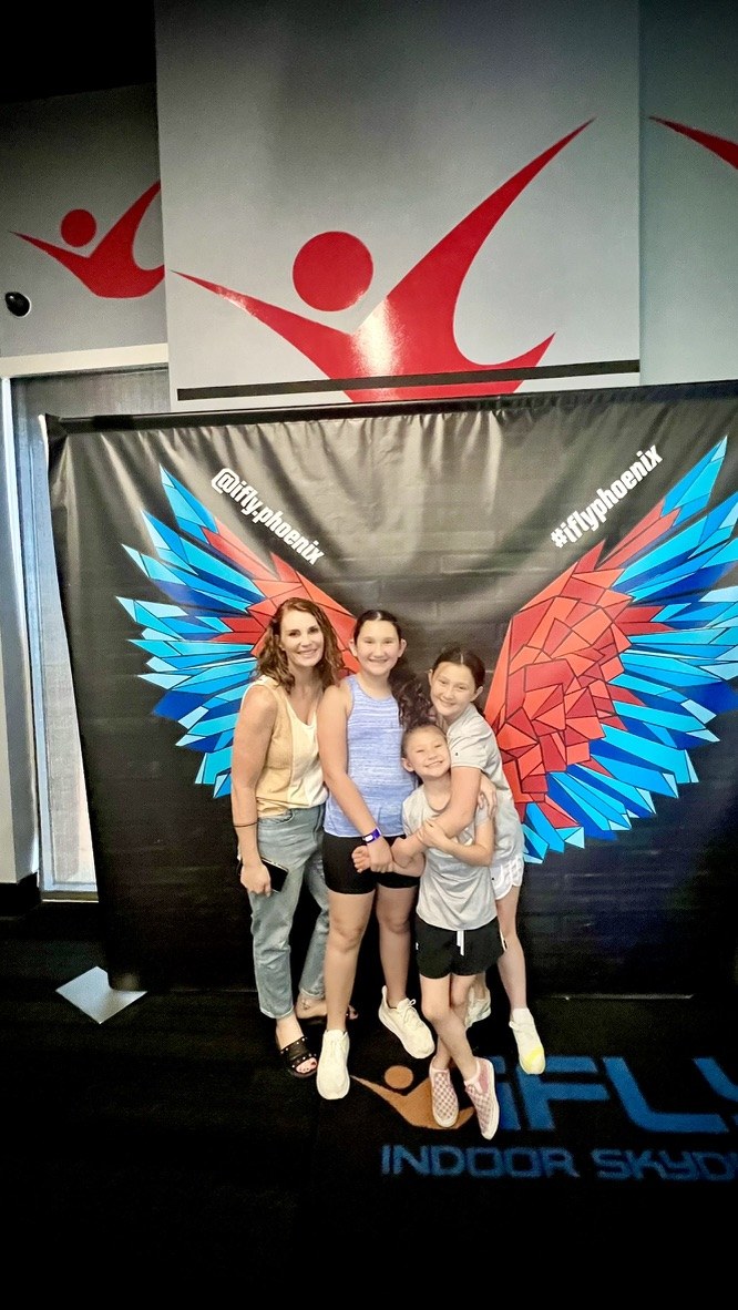 Ashley So with her three daughters at iFLY Phoenix, standing together in front of the winged mural.