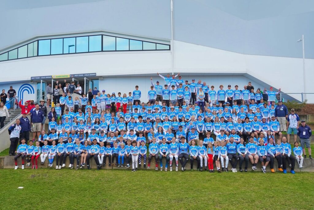 Team USA BMX riders of all ages in matching blue USA jerseys, gathered on steps in front of an arena for a group photo