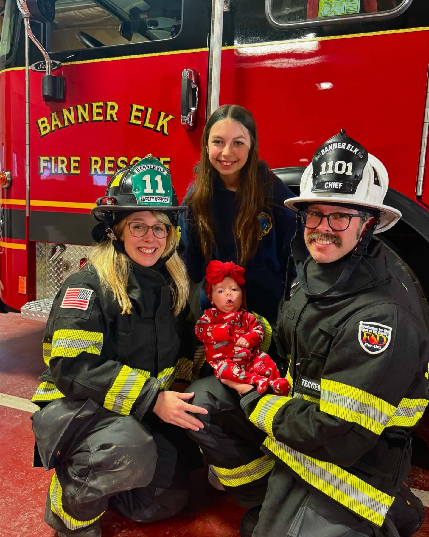 Tyler and Megan Burr in Banner Elk Fire Rescue turnout gear and helmets, kneeling beside a red fire engine with their two daughters, the youngest a baby in a red bow.