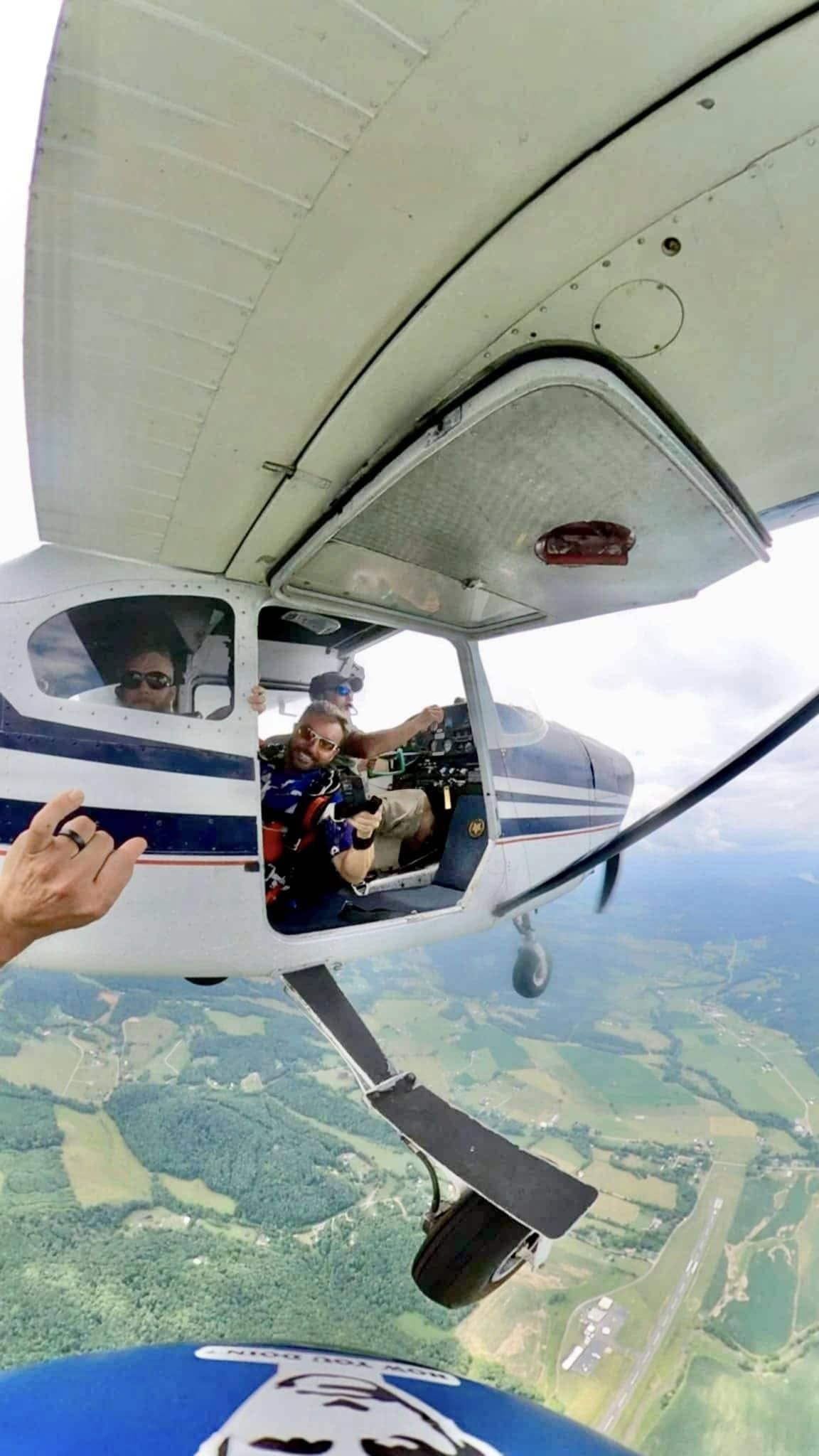 View from Tyler's helmet camera as he exits a small aircraft in flight: the pilot and jumpers are visible in the open door, with farmland and an airfield far below.