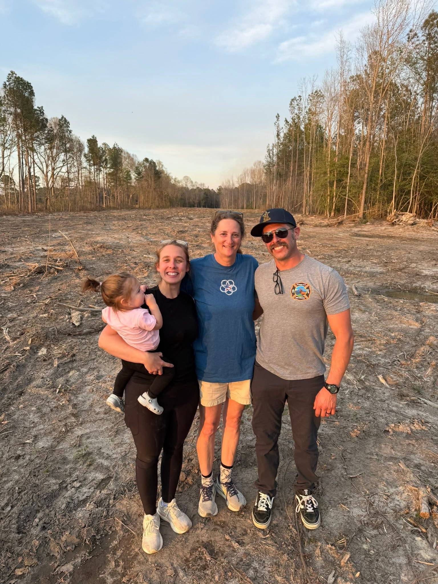 Linda Plummer, Megan Burr holding her young daughter, and Tyler Burr standing together on cleared land at Runway 37 on the CFH campus, with tall pines behind them.