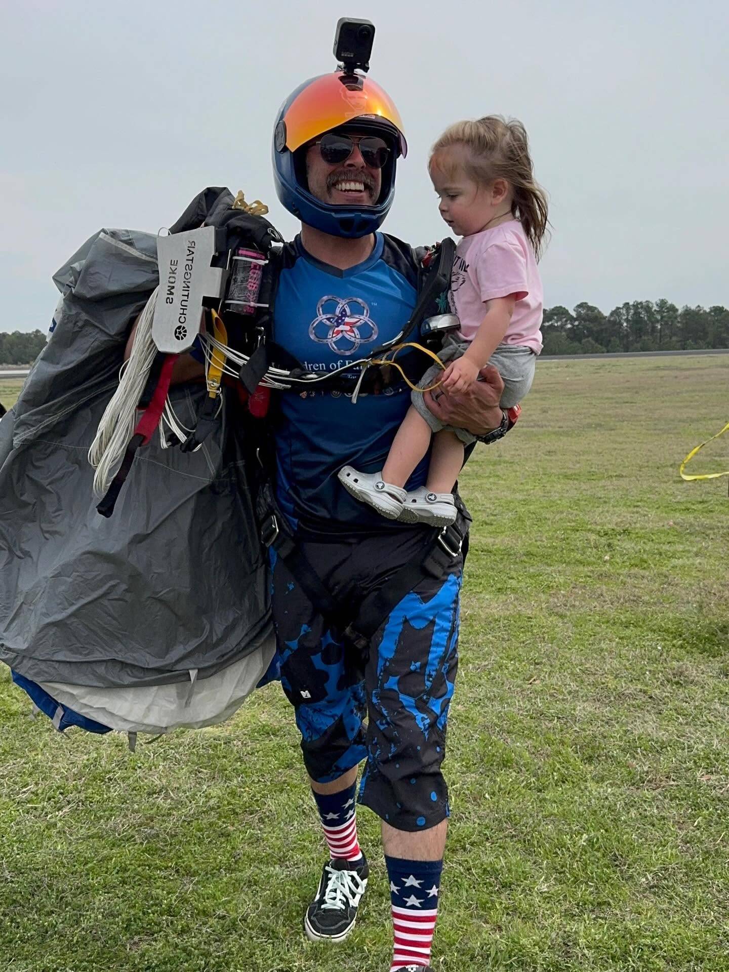 Tyler Burr, in a skydiving helmet and jump gear with his parachute gathered over one shoulder, carrying his young daughter across a grass landing area.