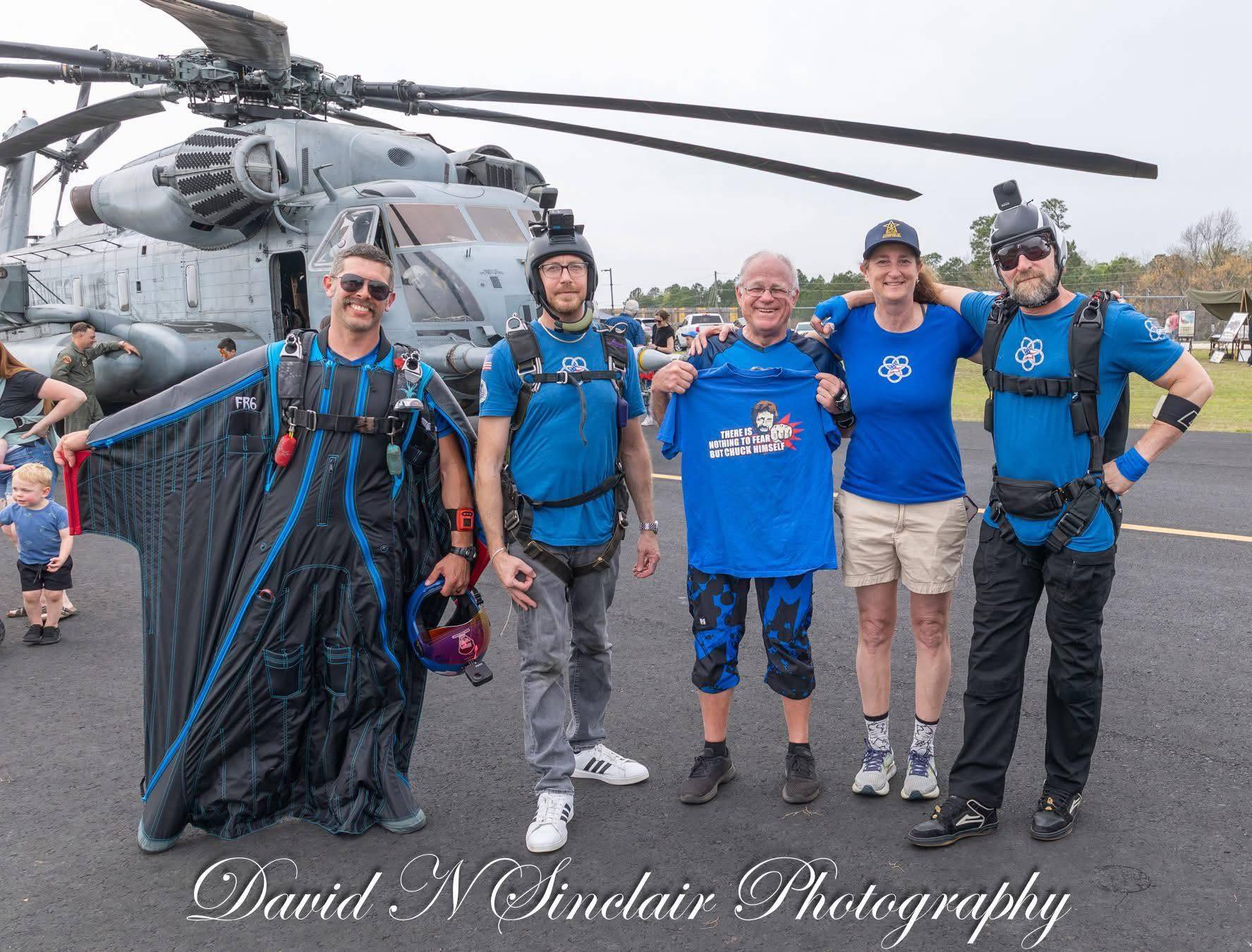 Tyler Burr in a black wingsuit standing with four other people on a tarmac in front of a large military helicopter.