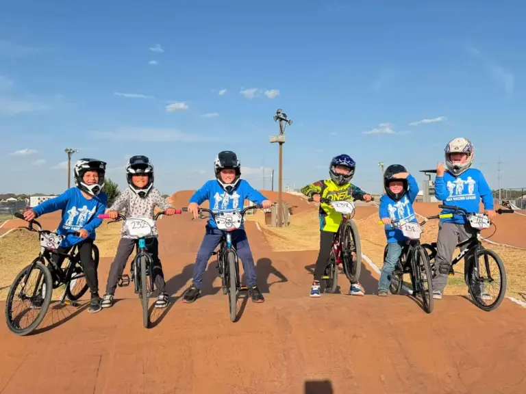 Six young BMX riders in matching jerseys with number plates lined up side by side on a BMX track