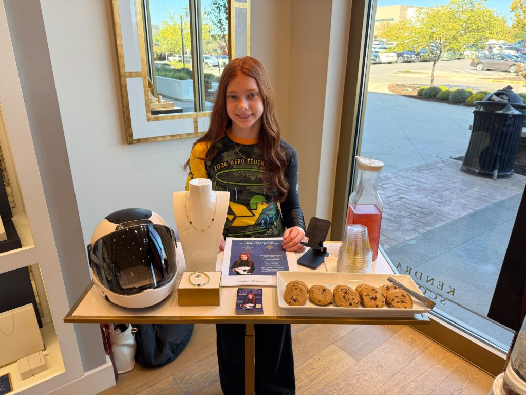 A CFH NEXT GEN athlete welcomes visitors beside jewelry and an indoor skydiving helmet at the Kendra Scott giveback event.